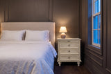 Osimo nightstand in Natural White in a grey panelled bedroom at dusk, a small lamp lit on top