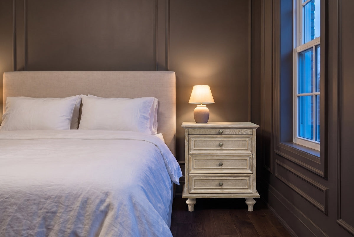 Osimo nightstand in Natural White in a grey panelled bedroom at dusk, a small lamp lit on top