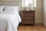 Osimo nightstand beside a bed with white linen against a limestone wall, a lamp and a stoneware bowl on top