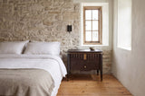 Pavia nightstand in Dark Brown beside a bed against a limestone wall in an old Quebec City house