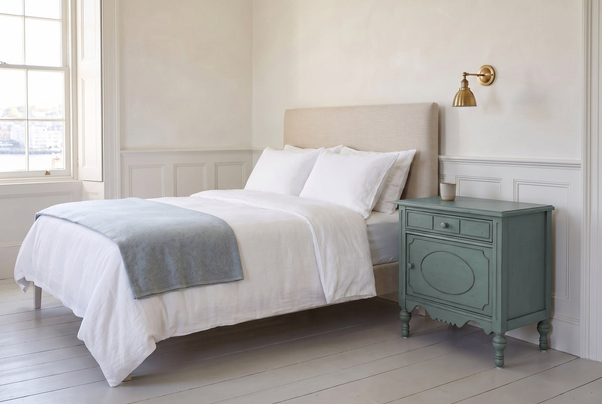 Perugia nightstand in Aged Blue beside a bed with white linen in a bright Halifax bedroom with white wainscot and a brass sco