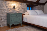 Perugia nightstand in Aged Blue against a fieldstone wall in a Quebec City bedroom at dusk, an iron sconce lit above it