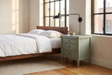 Pescara nightstand in Sage Green beside a walnut bed in a whitewashed brick Montreal loft, a black swing-arm lamp above it
