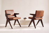 Two Pierre Jeanneret-style dining chairs in dark brown oak and cognac leather, one seen from the front and one from behind, with figs in a cream bowl on a travertine block between them