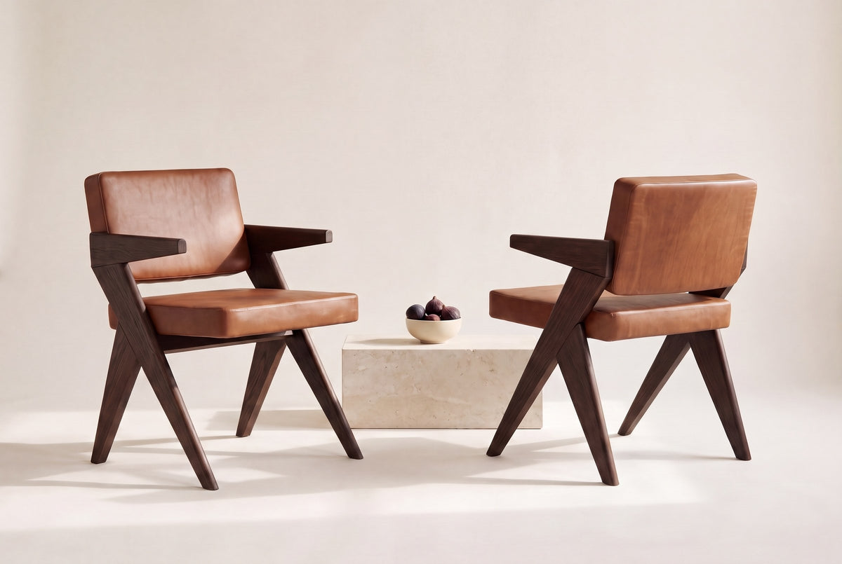 Two Pierre Jeanneret-style dining chairs in dark brown oak and cognac leather, one seen from the front and one from behind, with figs in a cream bowl on a travertine block between them