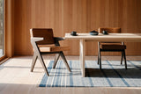 Two Pierre Jeanneret-style dining chairs in dark brown oak and cognac leather at a pale ash table with two black coffee cups and a bowl of blackberries, on a blue and ivory striped rug, against vertical cedar board walls beside a tall window in morning sun