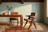Two Pierre Jeanneret-style dining chairs in dark brown oak and cognac leather at a cherry wood table with a bowl of pears and a pale blue vase holding a leafy branch, on a rust, ochre and cream rug over a concrete floor, against sea-green painted boards under a dark wood beam in golden evening light
