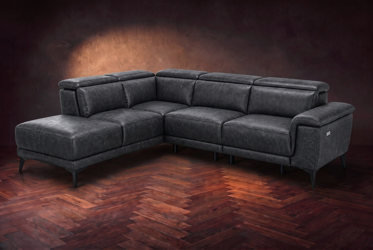 The Pista sectional in Aged Slate distressed full leather on a dark walnut herringbone floor against a brown plaster wall, three-quarter view from the arm end, headrests down and footrest closed. Shown in Aged Slate, Left chaise.