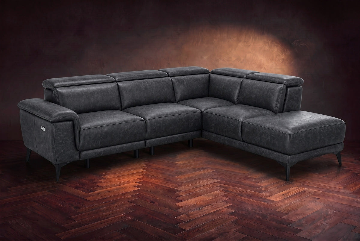 The Pista sectional in Aged Slate distressed full leather on a dark walnut herringbone floor against a brown plaster wall, three-quarter view from the arm end, headrests down and footrest closed. Shown in Aged Slate, Right chaise.