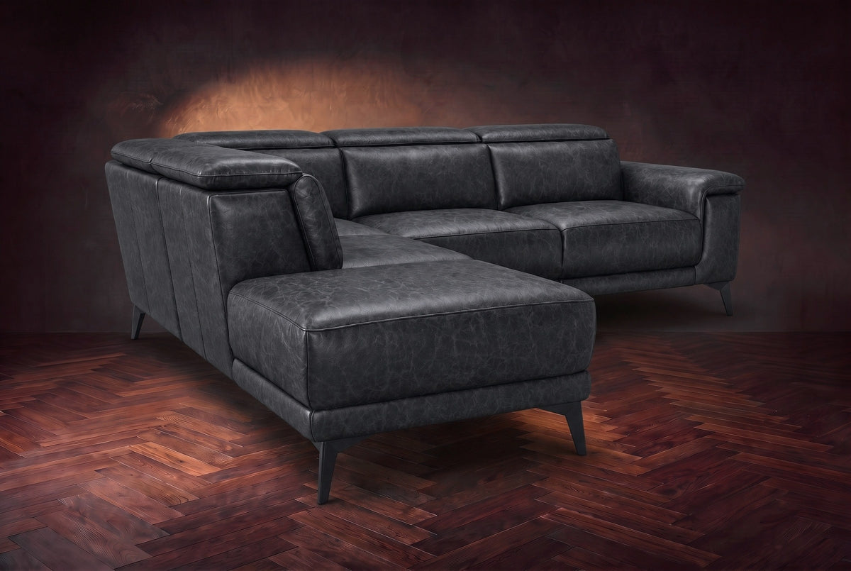 The Pista sectional in Aged Slate distressed full leather on a dark walnut herringbone floor against a brown plaster wall, seen from the chaise end, headrests down and footrest closed. Shown in Aged Slate, Left chaise.