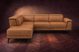 The Pista sectional in Antiqued Caramel distressed full leather on a dark walnut herringbone floor against a brown plaster wall, seen from the front with the chaise toward the camera, headrests down and footrest closed. Shown in Antiqued Caramel, Left chaise.