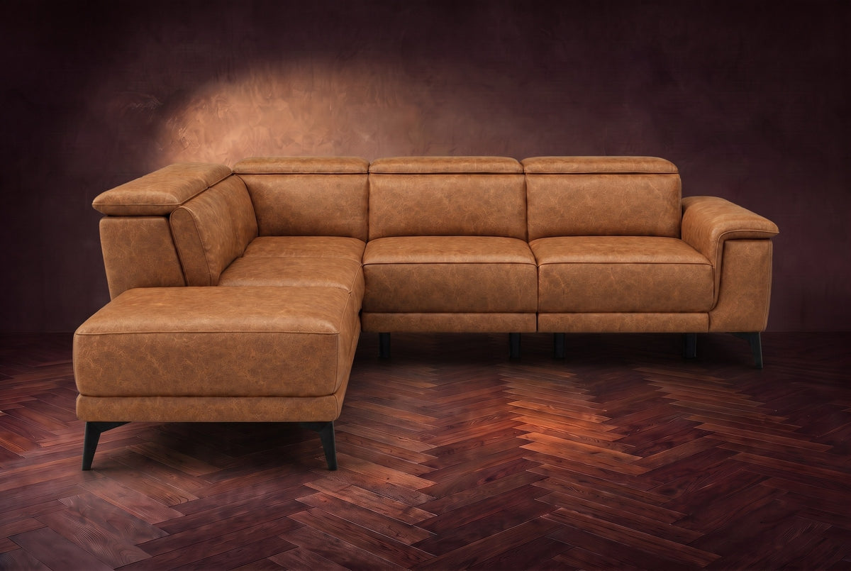 The Pista sectional in Antiqued Caramel distressed full leather on a dark walnut herringbone floor against a brown plaster wall, seen from the front with the chaise toward the camera, headrests down and footrest closed. Shown in Antiqued Caramel, Left chaise.