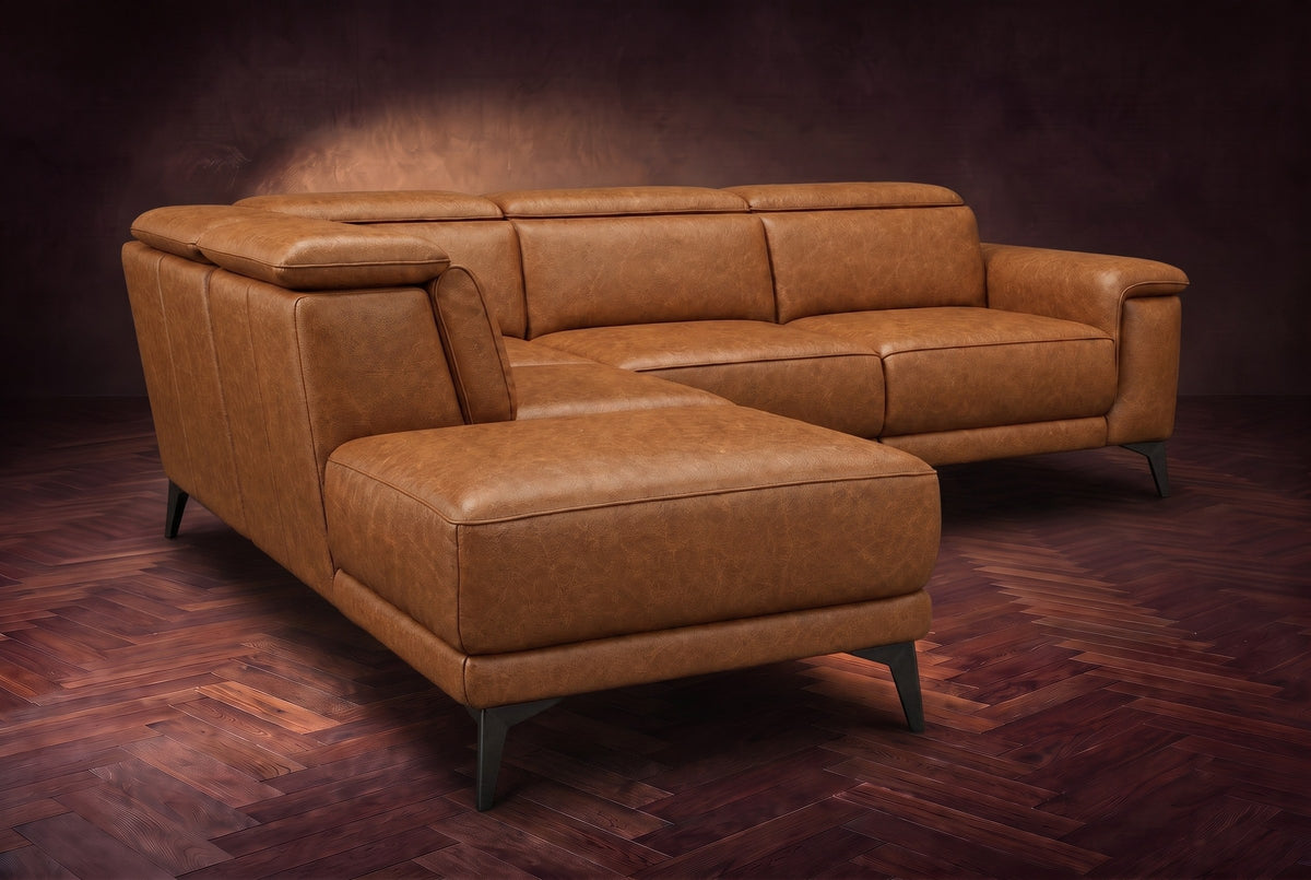 The Pista sectional in Antiqued Caramel distressed full leather on a dark walnut herringbone floor against a brown plaster wall, seen from the chaise end, headrests down and footrest closed. Shown in Antiqued Caramel, Left chaise.