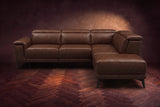 The Pista sectional in Burnished Chestnut distressed full leather on a dark walnut herringbone floor against a brown plaster wall, seen from the front with the chaise toward the camera, headrests down and footrest closed. Shown in Burnished Chestnut, Right chaise.