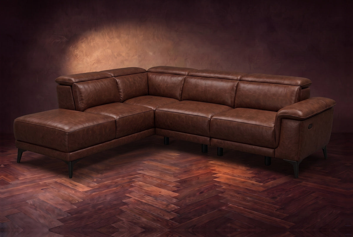 The Pista sectional in Burnished Chestnut distressed full leather on a dark walnut herringbone floor against a brown plaster wall, three-quarter view from the arm end, headrests down and footrest closed. Shown in Burnished Chestnut, Left chaise.