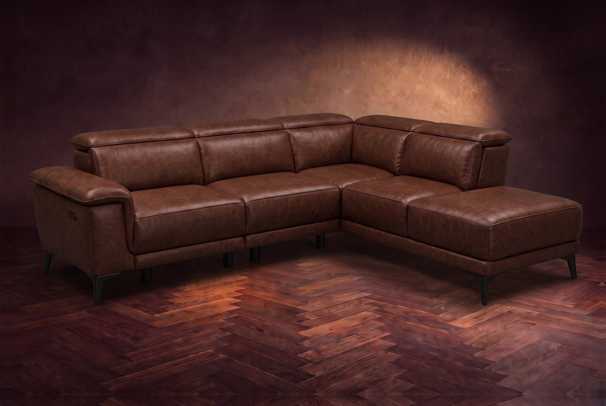 The Pista sectional in Burnished Chestnut distressed full leather on a dark walnut herringbone floor against a brown plaster wall, three-quarter view from the arm end, headrests down and footrest closed. Shown in Burnished Chestnut, Right chaise.