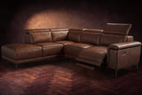 The Pista sectional in Burnished Chestnut distressed full leather on a dark walnut herringbone floor against a brown plaster wall, the seat beside the box arm reclined with its two-tier footrest out and its headrest raised, the other headrests down. Shown in Burnished Chestnut, Left chaise.