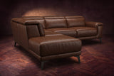 The Pista sectional in Burnished Chestnut distressed full leather on a dark walnut herringbone floor against a brown plaster wall, seen from the chaise end, headrests down and footrest closed. Shown in Burnished Chestnut, Left chaise.