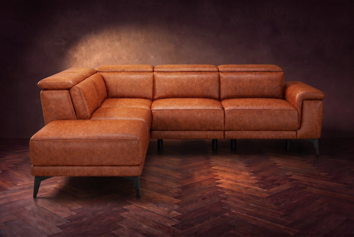 The Pista sectional in Tuscan Cognac distressed full leather on a dark walnut herringbone floor against a brown plaster wall, seen from the front with the chaise toward the camera, headrests down and footrest closed. Shown in Tuscan Cognac, Left chaise.