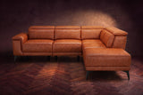 The Pista sectional in Tuscan Cognac distressed full leather on a dark walnut herringbone floor against a brown plaster wall, seen from the front with the chaise toward the camera, headrests down and footrest closed. Shown in Tuscan Cognac, Right chaise.