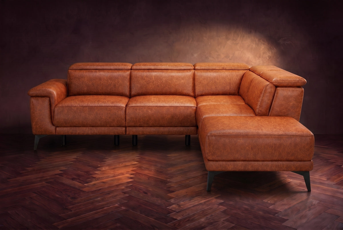 The Pista sectional in Tuscan Cognac distressed full leather on a dark walnut herringbone floor against a brown plaster wall, seen from the front with the chaise toward the camera, headrests down and footrest closed. Shown in Tuscan Cognac, Right chaise.