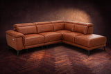 The Pista sectional in Tuscan Cognac distressed full leather on a dark walnut herringbone floor against a brown plaster wall, three-quarter view from the arm end, headrests down and footrest closed. Shown in Tuscan Cognac, Right chaise.