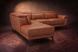 The Pista sectional in Tuscan Cognac distressed full leather on a dark walnut herringbone floor against a brown plaster wall, seen from the chaise end, headrests down and footrest closed. Shown in Tuscan Cognac, Left chaise.