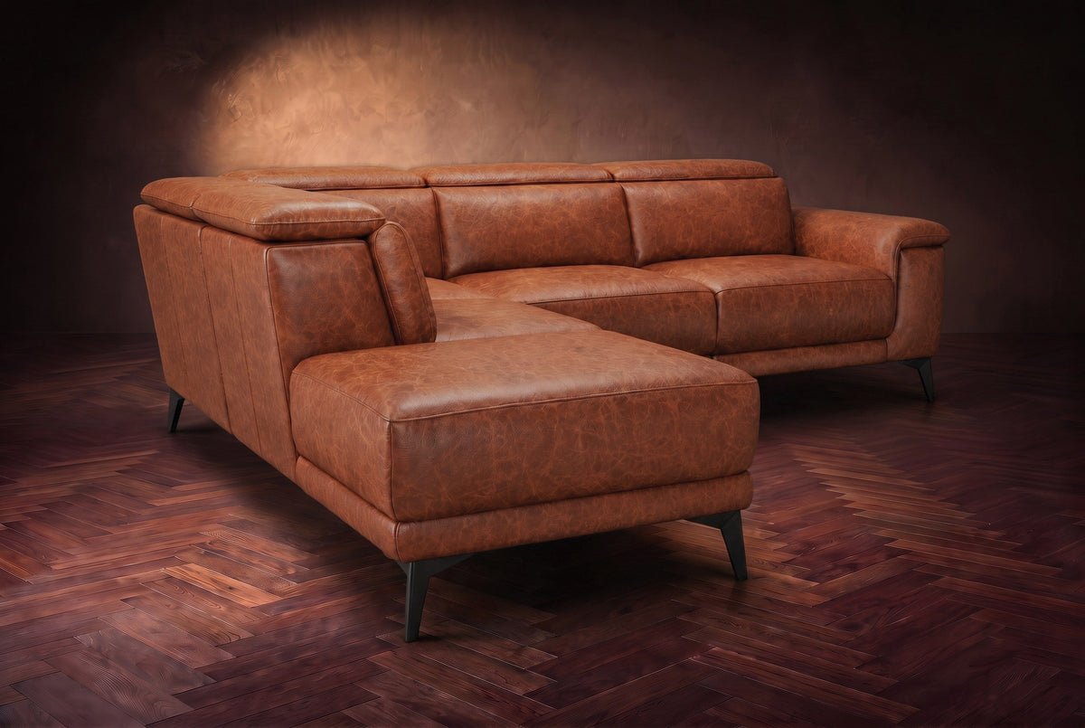 The Pista sectional in Tuscan Cognac distressed full leather on a dark walnut herringbone floor against a brown plaster wall, seen from the chaise end, headrests down and footrest closed. Shown in Tuscan Cognac, Left chaise.