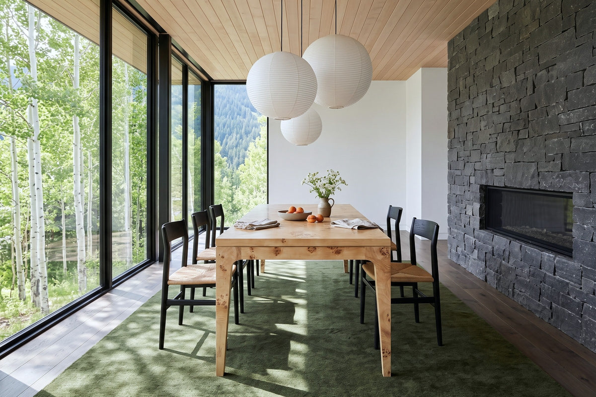 End view of the Polcenigo dining table in Honey Blonde Burl in a Rossland mountain house, with a dark stone fireplace wall