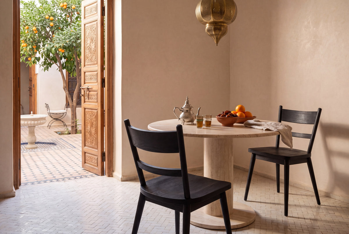 Two black Rocco ladder-back dining chairs at a table with mint tea under a brass lantern in a restored Marrakech riad, carved cedar doors open to an orange-tree courtyard with a fountain