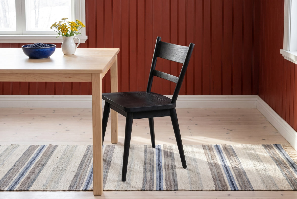 One black wire-brushed oak two-slat Rocco dining chair at the end of a pale birch table with a blue bowl of blueberries and a white jug of yellow flowers, in a red board-walled room with white trim, a pale pine floor and a striped rag rug