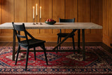 Two black wire-brushed oak two-slat Rocco dining chairs at a long pale stone table on a dark bronze trestle base, with three lit candles in brass candlesticks and a green bowl of red pears, in a honey oak panelled room with a red and navy patterned rug