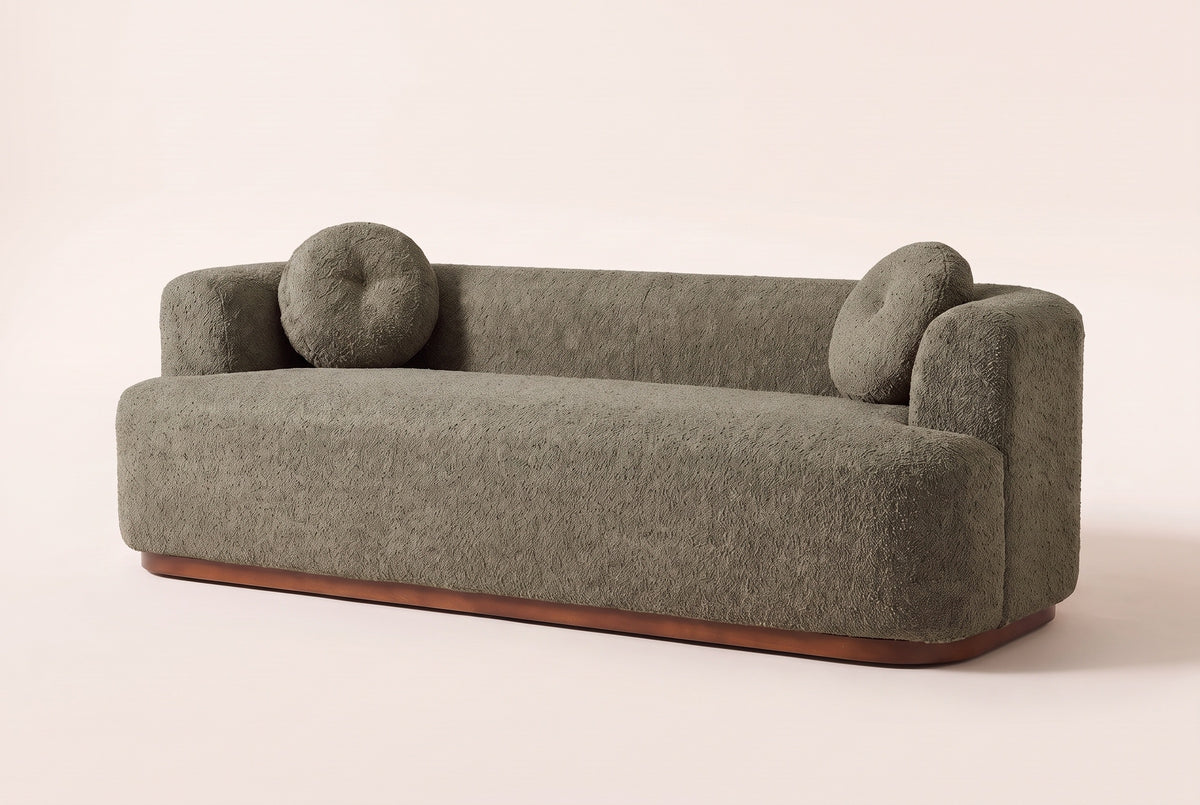 Sabbioneta boucle sofa in olive green on a walnut-brown wooden plinth, three-quarter view on a warm off-white studio backdrop