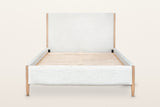 Sarnano bed, front view from the foot on a warm white studio backdrop: the upholstered foot rail between two round oak posts
