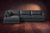 The Serena sectional in Aged Slate distressed full leather on a walnut herringbone floor against a dark plaster wall, seen from the front. Shown in Aged Slate, Left bumper.