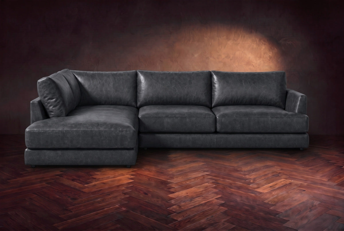 The Serena sectional in Aged Slate distressed full leather on a walnut herringbone floor against a dark plaster wall, seen from the front. Shown in Aged Slate, Left bumper.