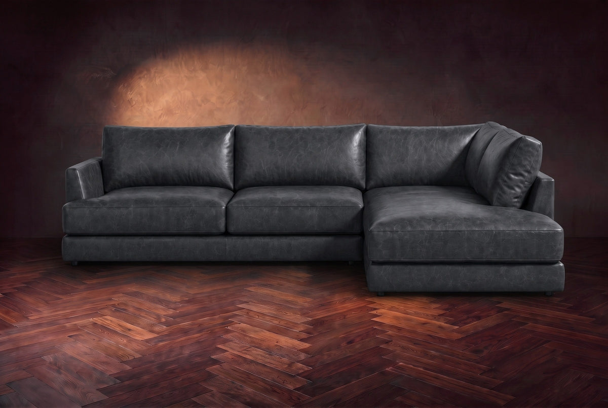 The Serena sectional in Aged Slate distressed full leather on a walnut herringbone floor against a dark plaster wall, seen from the front. Shown in Aged Slate, Right bumper.