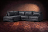 The Serena sectional in Aged Slate distressed full leather on a walnut herringbone floor against a dark plaster wall, three-quarter view. Shown in Aged Slate, Left bumper.