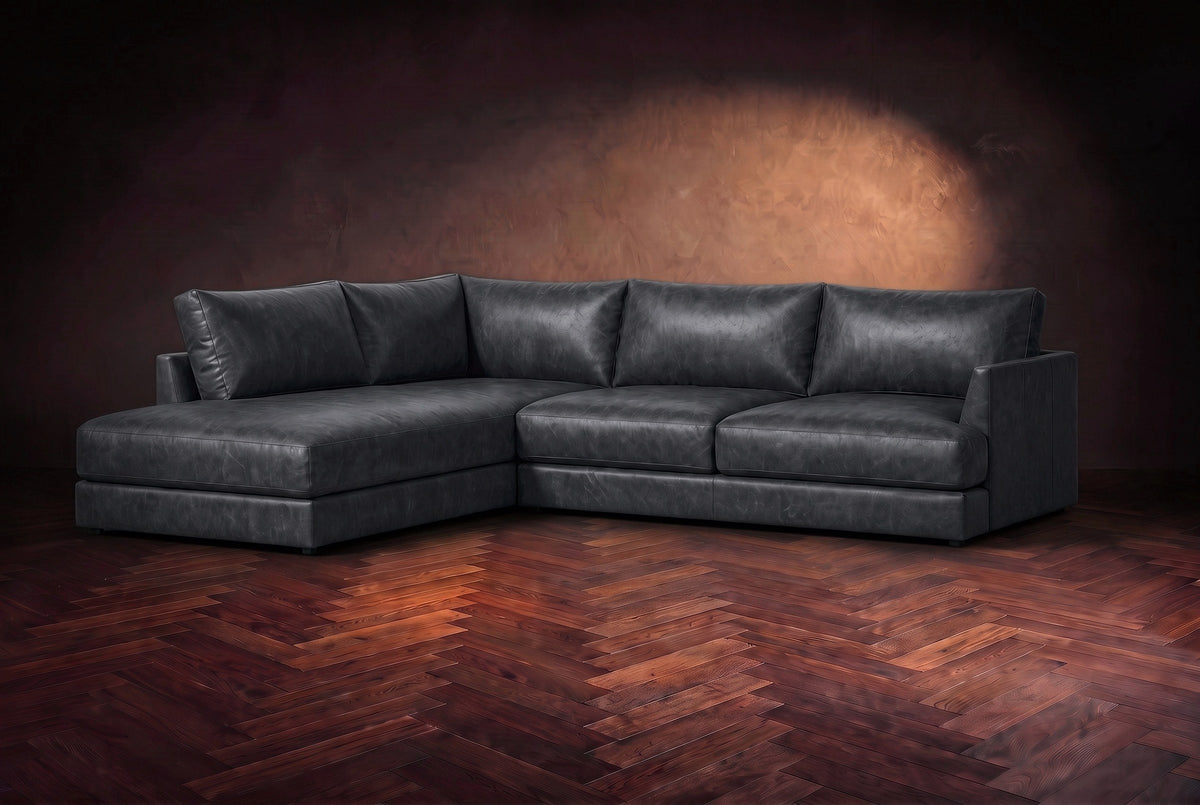 The Serena sectional in Aged Slate distressed full leather on a walnut herringbone floor against a dark plaster wall, three-quarter view. Shown in Aged Slate, Left bumper.