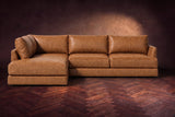 The Serena sectional in Antiqued Caramel distressed full leather on a walnut herringbone floor against a dark plaster wall, seen from the front. Shown in Antiqued Caramel, Left bumper.