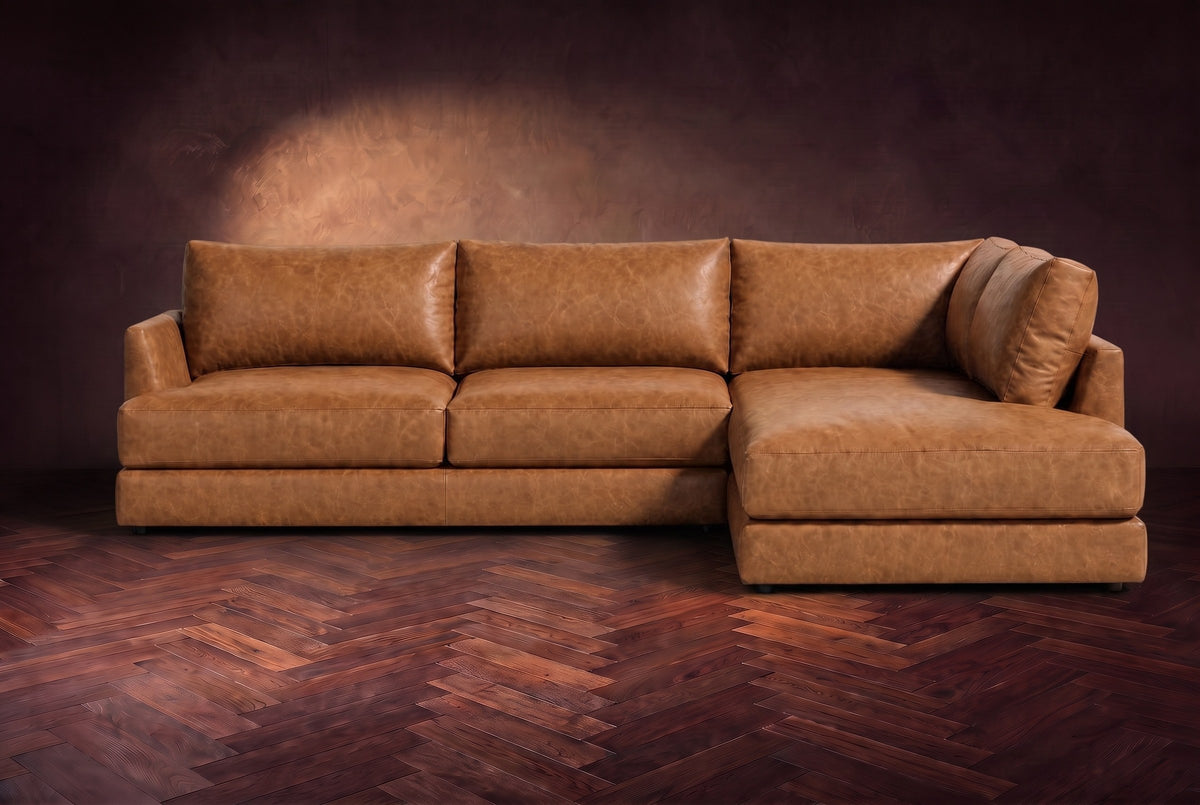 The Serena sectional in Antiqued Caramel distressed full leather on a walnut herringbone floor against a dark plaster wall, seen from the front. Shown in Antiqued Caramel, Right bumper.