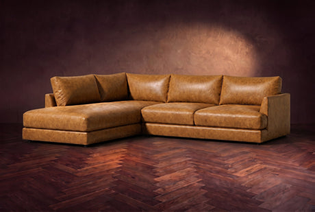 The Serena sectional in Antiqued Caramel distressed full leather on a walnut herringbone floor against a dark plaster wall, three-quarter view. Shown in Antiqued Caramel, Left bumper.
