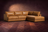The Serena sectional in Antiqued Caramel distressed full leather on a walnut herringbone floor against a dark plaster wall, three-quarter view. Shown in Antiqued Caramel, Right bumper.