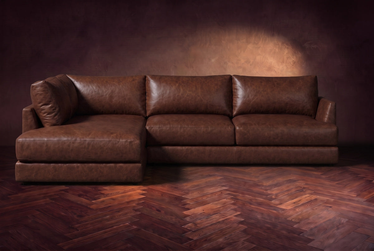 The Serena sectional in Burnished Chestnut distressed full leather on a walnut herringbone floor against a dark plaster wall, seen from the front. Shown in Burnished Chestnut, Left bumper.