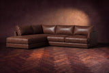 The Serena sectional in Burnished Chestnut distressed full leather on a walnut herringbone floor against a dark plaster wall, three-quarter view. Shown in Burnished Chestnut, Left bumper.