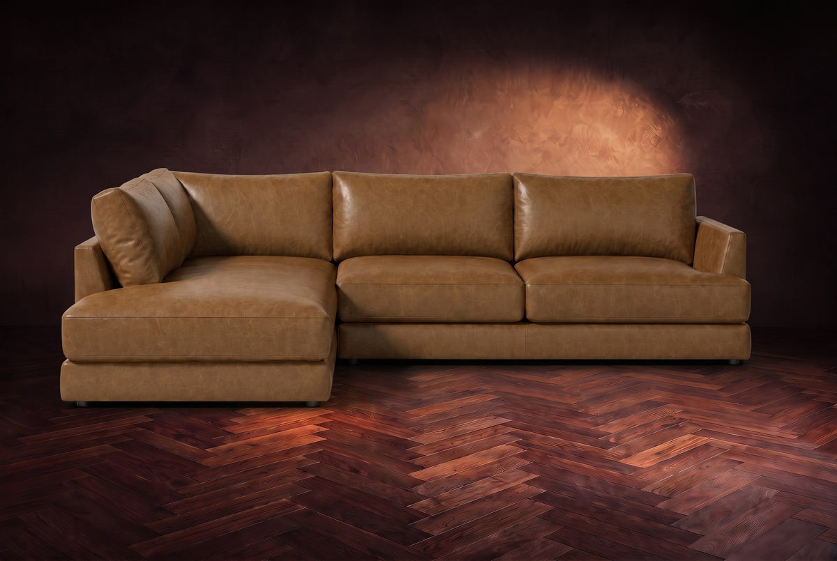 The Serena sectional in Weathered Saddle distressed full leather on a walnut herringbone floor against a dark plaster wall, seen from the front. Shown in Weathered Saddle, Left bumper.