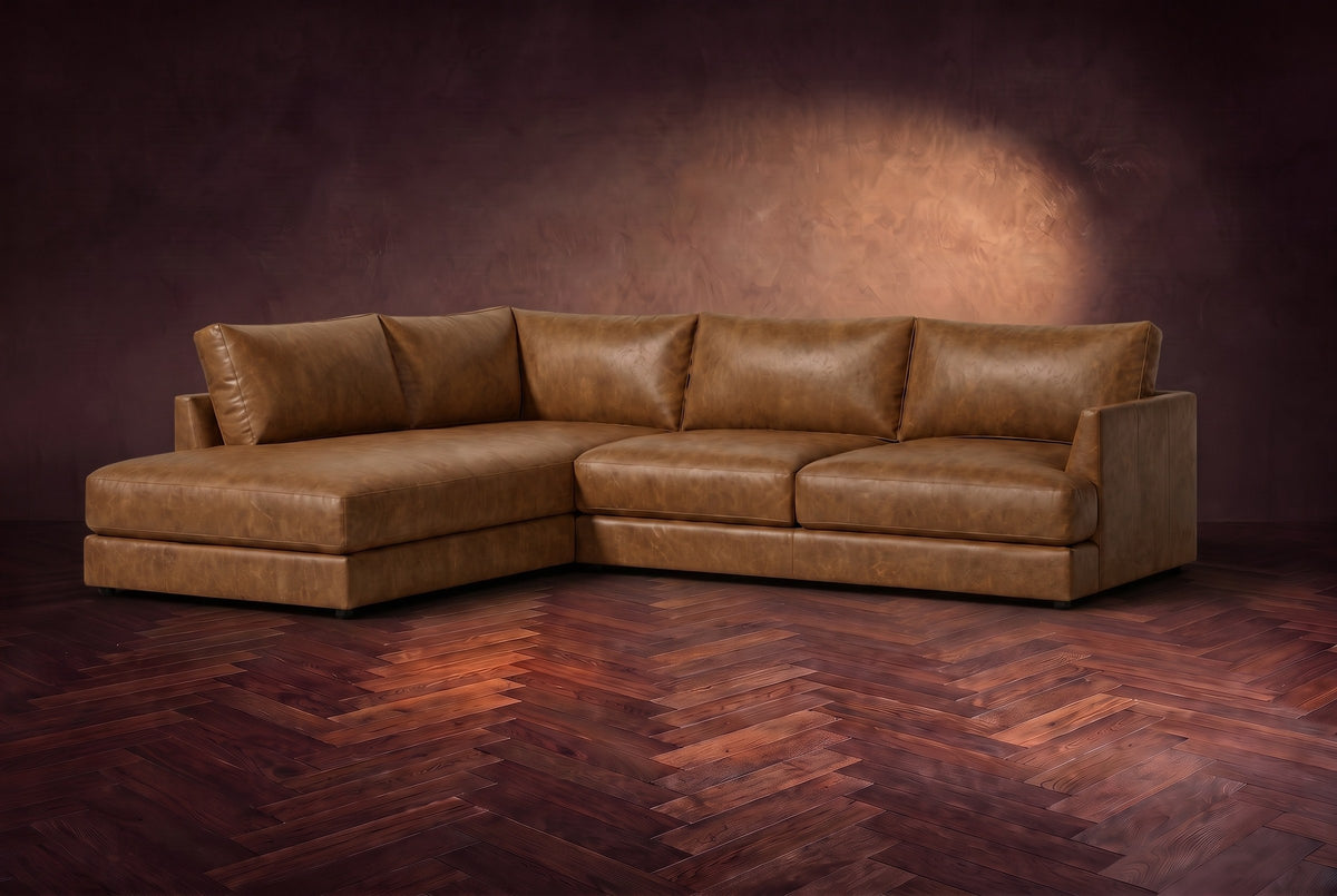 The Serena sectional in Weathered Saddle distressed full leather on a walnut herringbone floor against a dark plaster wall, three-quarter view. Shown in Weathered Saddle, Left bumper.