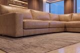 Low view along the Serena corner sectional from its arm end toward the corner, on a rug in a room with lit shelving and blue-hour windows. Shown in Washed Sand.