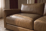 Close-up of the Serena corner sectional's slim track arm running the full depth of the seat, beside sheer linen curtains on a terrazzo floor. Shown in Weathered Saddle.