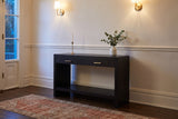 Sibari console table in a plaster entry hall in Victoria at dusk, brass sconces lit, a candlestick and eucalyptus on top
